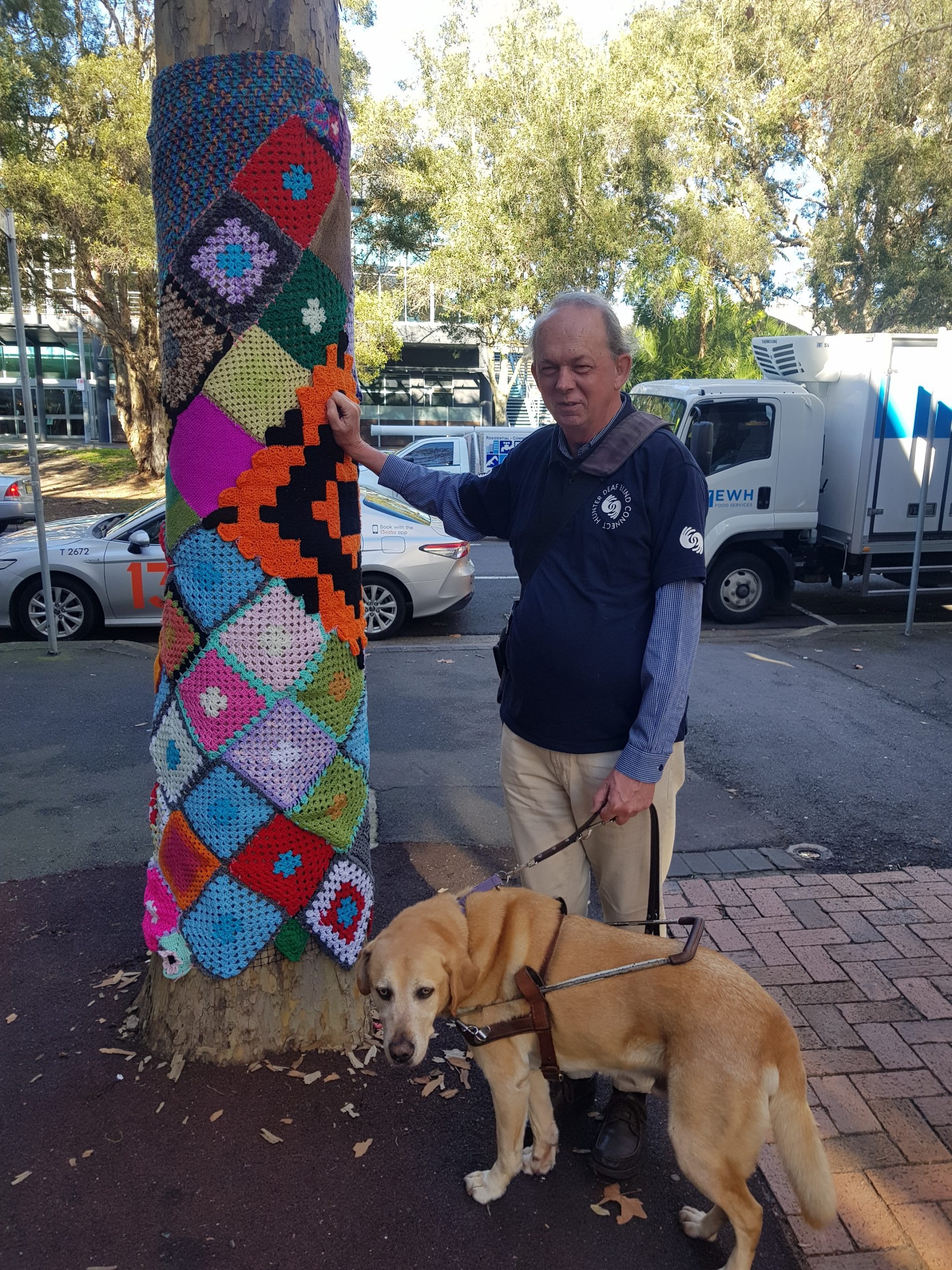 Stephen standing next to tree that has been covered in colourful pieces of knitted wool. Next to Stephen his guide dog