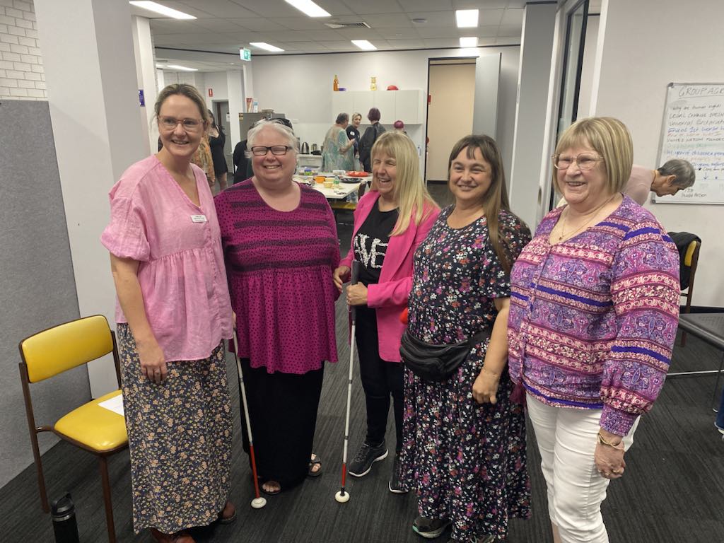 Image description: Five woman in multi-coloured clothes standing with their arms around one another smiling